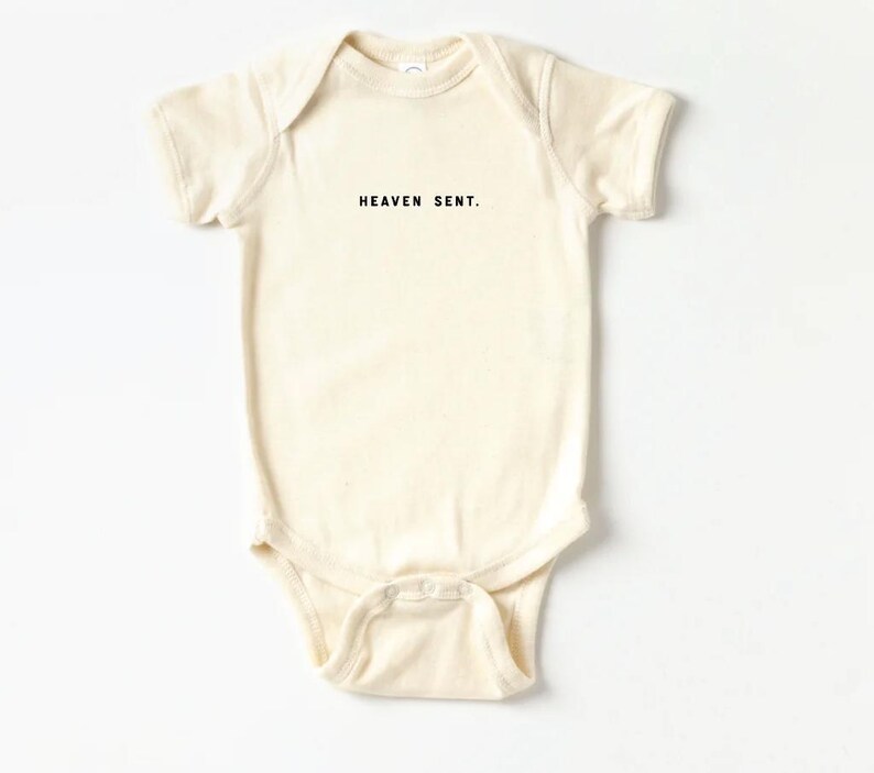 Op de afbeelding: Een cr&egrave;mekleurige baby romper met korte mouwen en de tekst "HEAVEN SENT." in het zwart gedrukt.