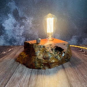 May include: A rustic wooden lamp base with a warm, glowing Edison bulb. The base is made of natural wood with a rough, textured surface. The lamp is lit and casting a warm glow.