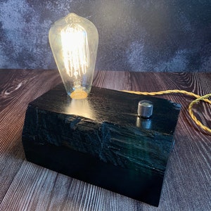 May include: A black wooden lamp base with a vintage-style light bulb and a silver toggle switch. The lamp is on a wooden surface.