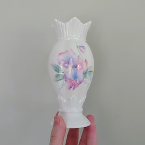 Può includere: Un vaso in ceramica bianca con un disegno floreale con fiori rosa, blu e verdi. Il vaso ha un bordo a festoni e una base svasata.