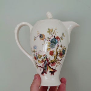 Puede incluir: Tetera de cerámica blanca con un diseño floral que presenta un árbol con pájaros y flores en tonos rojos, amarillos, azules y verdes.
