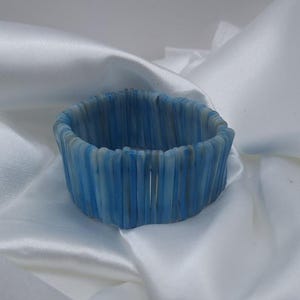 Pode incluir: Uma pulseira elástica composta por numerosos segmentos verticais, finos e cilíndricos em vários tons de azul, azul claro e branco. Os segmentos estão dispostos lado a lado para formar uma estrutura de banda circular flexível.