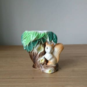 May include: A ceramic planter shaped like a tree trunk with green leaves and a squirrel climbing up the side. The squirrel is brown and white with a bushy tail.