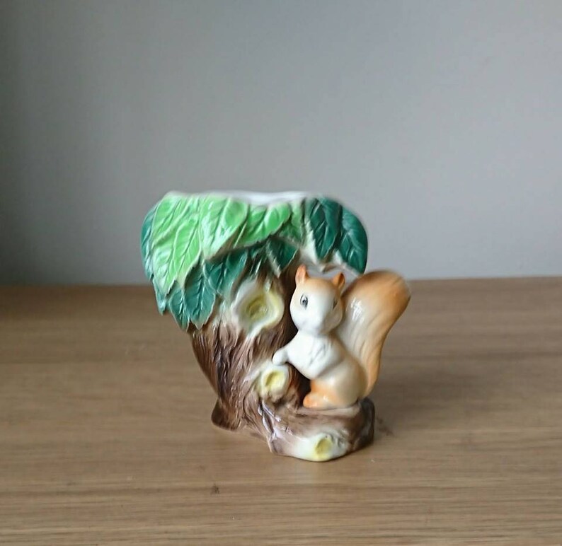 May include: A ceramic planter shaped like a tree trunk with green leaves and a squirrel climbing up the side. The squirrel is brown and white with a bushy tail.