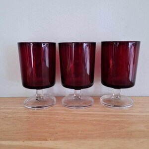 May include: Three clear glass wine goblets with a deep red colored base. The goblets have a short stem and a round base.