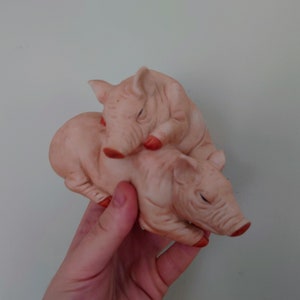 Puede incluir: Dos figuritas de cerdos de cerámica, una encima de la otra, pintadas de rosa y rojo. El cerdo de arriba está acostado con la cabeza apoyada en la espalda del otro cerdo.