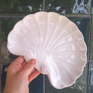 Può includere: Un piatto in ceramica bianco lucido a forma di grande conchiglia. Il piatto presenta un bordo smerlato e profondi solchi radianti che si estendono dal centro verso il bordo esterno, creando un aspetto strutturato a forma di ventaglio.