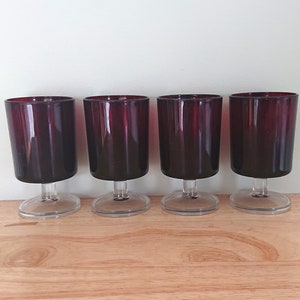 Puede incluir: Cuatro copas de vino con tallo de vidrio transparente con un tinte rojo oscuro. Las copas están dispuestas en una fila sobre una superficie de madera.