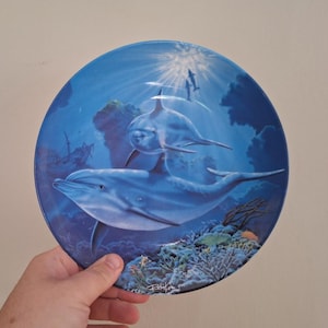 Puede incluir: Plato decorativo con dos delfines nadando en un océano azul. El plato tiene un acabado brillante e incluye un naufragio y detalles de arrecifes de coral. El diseño evoca la profundidad del mar.