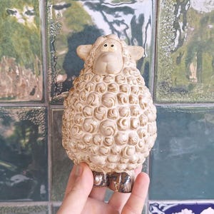 May include: A ceramic sheep figurine featuring a textured, cream-colored wool body with brown highlights. The sheep has a smooth, light-colored face with two small eyes, rounded ears, and dark brown hooves at the base of its rounded, plump body.