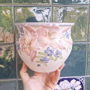Könnte beinhalten: Ein runder Keramikübertopf mit cremefarbener Basis und erhabenen, dreidimensionalen Blumenreliefmustern. Das Design umfasst große rosa Blüten und kleinere blaue Blumen mit grünen Blättern. Der Rand weist eine geflochtene Seilstruktur auf.