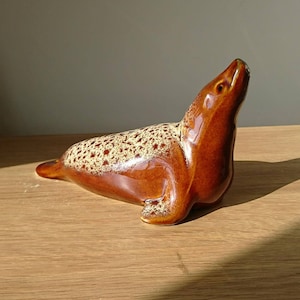 May include: A brown ceramic seal figurine with a speckled brown and cream glaze. The seal is posed with its head tilted up and its flippers outstretched.