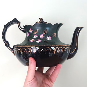 May include: A black teapot with a green and gold floral design. The teapot has a wide, round body and a curved spout. The handle is attached to the side of the teapot.
