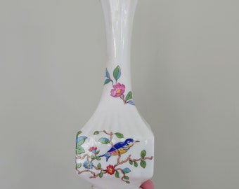 Puede incluir: Un jarrón de cerámica blanca con un borde dorado y un diseño floral con un pájaro azul, flores rosas y hojas verdes.