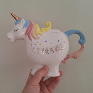 May include: A white ceramic unicorn teapot with a gold horn, rainbow mane, and blue handle. The teapot has the phrase "BELIEVE IN MAGIC" in gold lettering and is decorated with stars and crescent moons.