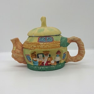 Puede incluir: Una tetera de cerámica amarilla y verde con forma de casa con un techo marrón y un asa marrón. La tetera está decorada con cuatro osos que llevan ropa y una ventana con cristales azules y blancos.