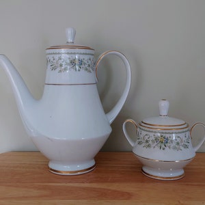 Może przedstawiać: Biały porcelanowy dzbanek do kawy i cukiernica z kwiatowym wzorem i złotą obwódką.