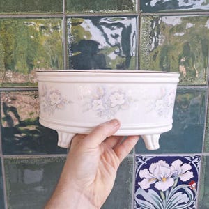 May include: A round, off-white ceramic planter featuring a delicate, repeating floral pattern in soft pastel shades of blue and green. The vessel is supported by three short, curved feet and has a smooth, glossy finish with a simple rim design.