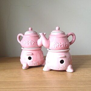 May include: Two pink ceramic teapots with floral designs, each sitting on a matching pink stand with three legs.