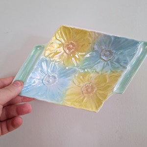 Puede incluir: Una bandeja decorativa de cerámica con un diseño floral. La bandeja es rectangular con forma de diamante y presenta cuatro flores en relieve en amarillo y azul. La bandeja tiene un acabado brillante y está sostenida por una mano.