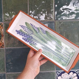 Könnte beinhalten: Rechteckige Servierplatte aus Keramik mit weißem Innenraum, terrakottafarbenem Rand und Griffen. Die Platte hat ein handgemaltes Design aus Lavendelzweigen in Grün- und Lilatönen. Die Platte wird von einer Person gehalten.