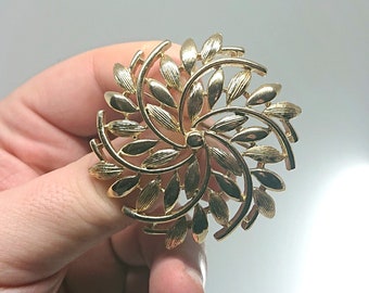May include: A gold-toned brooch with a swirling design of stylized leaves. The brooch is a delicate and intricate piece of jewelry.