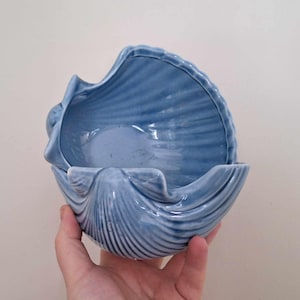 Può includere: Una ciotola a forma di conchiglia in ceramica blu chiaro. La ciotola ha una superficie strutturata e scanalata, e l'interno è liscio. La ciotola è tenuta in una mano. La ciotola è probabilmente utilizzata per scopi decorativi o come piccolo piatto.