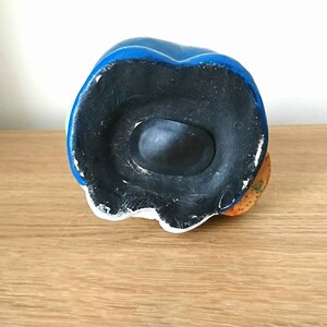 May include: The bottom of a ceramic piggy bank shaped like a person wearing a blue hat and a yellow shirt. The piggy bank has a black rubber stopper on the bottom.