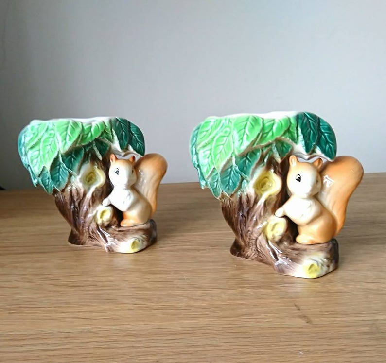 May include: Two ceramic squirrel planter vases with green leaves and brown tree trunks. The squirrels are brown and white with black eyes.