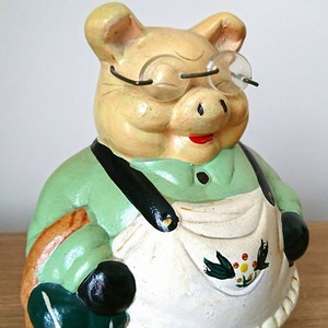 May include: A green and white ceramic piggy bank with a red mouth and glasses. The piggy bank is wearing a green shirt and a white apron with a floral design.