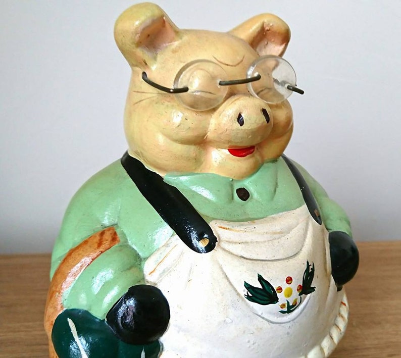 May include: A green and white ceramic piggy bank with a red mouth and glasses. The piggy bank is wearing a green shirt and a white apron with a floral design.