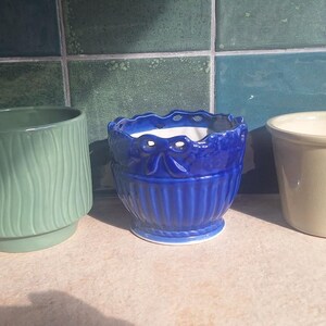 Può includere: Tre vasi per piante in ceramica disposti in fila. Il vaso a sinistra è verde salvia con onde verticali. Il vaso centrale è blu intenso con bordo smerlato e scanalature verticali. Il vaso a destra è color crema con un piccolo dettaglio floreale in rilievo.