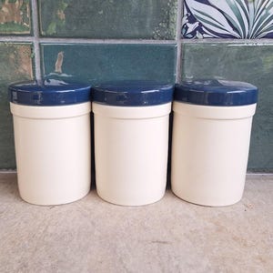 May include: Three cylindrical storage canisters with smooth off-white bodies and glossy dark blue lids. Each container features a simple, uniform design with a straight vertical profile and a flat, circular top, arranged side by side in a row.