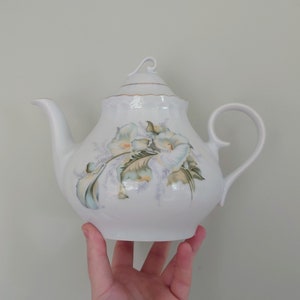 May include: White porcelain teapot with a floral design featuring white flowers with green leaves and a gold trim around the lid.