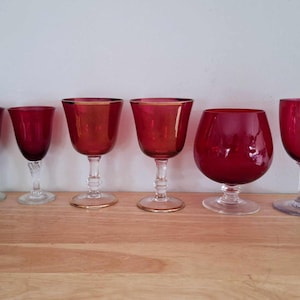 May include: Six red wine glasses of different shapes and sizes. The glasses are arranged in a row on a wooden surface.