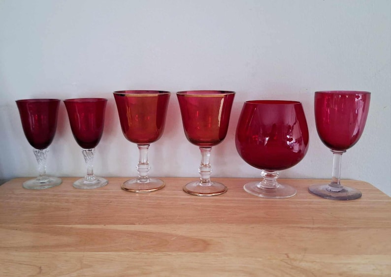May include: Six red wine glasses of different shapes and sizes. The glasses are arranged in a row on a wooden surface.