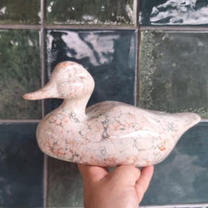 Könnte beinhalten: Keramik-Entenfigur mit einem gesprenkelten Muster in Creme, Orange und Grün. Die Ente hat einen langen, gebogenen Hals und einen kleinen, spitzen Schnabel. Die Figur wird in einer Hand vor einem Hintergrund aus grünen und türkisfarbenen Fliesen gehalten.