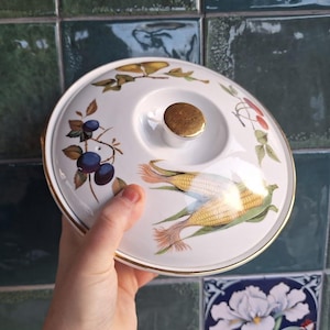 May include: A white ceramic serving dish lid with a gold rim and a gold knob. The lid is decorated with illustrations of corn, berries, and leaves. The lid is being held by a person.