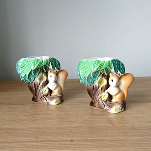 May include: A pair of ceramic squirrel candle holders. Each holder features a squirrel perched on a tree trunk with green leaves. The holders are white with brown and green accents.