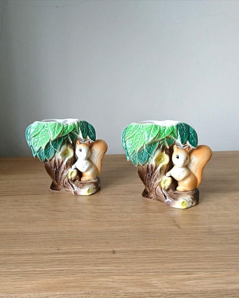 May include: A pair of ceramic squirrel candle holders. Each holder features a squirrel perched on a tree trunk with green leaves. The holders are white with brown and green accents.