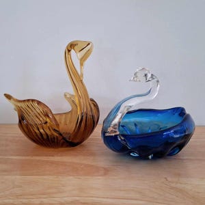 May include: Two glass swan-shaped dishes. One is amber glass and the other is blue glass with a clear glass swan head.