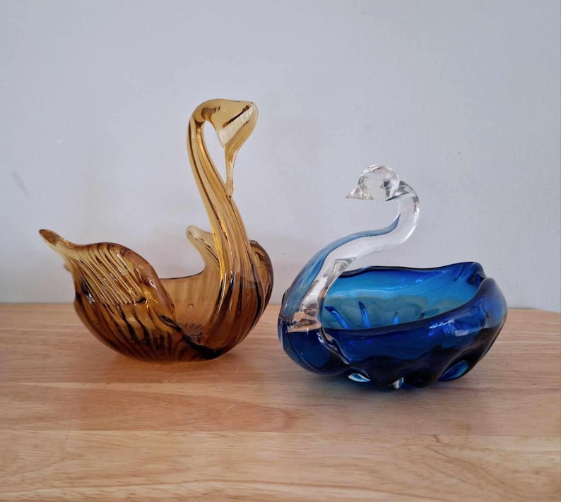 May include: Two glass swan-shaped dishes. One is amber glass and the other is blue glass with a clear glass swan head.