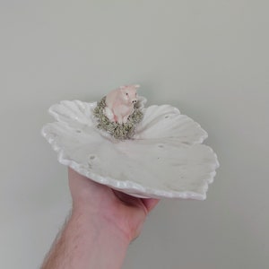 May include: A white ceramic leaf-shaped dish with a small pink pig figurine sitting on a bed of green moss in the centre.