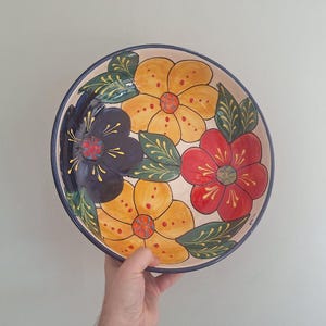 Può includere: Una ciotola rotonda in ceramica con base color crema caratterizzata da un motivo floreale dipinto a mano. Il design include due grandi fiori gialli, un fiore blu scuro e un fiore rosso, tutti accentati da foglie verdi e sottili linee gialle.