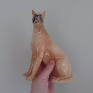 May include: A ceramic figurine of a tan-colored boxer dog sitting with its head tilted up. The dog has black eyes and a black nose.