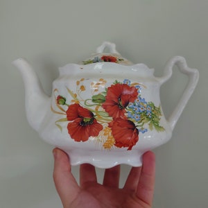 Pode incluir: Bule de chá branca com um design floral com papoulas vermelhas, miosotis azuis e espigas de trigo. A chaleira tem uma borda festonada e uma alça.