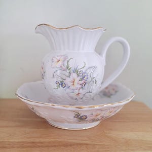 Könnte beinhalten: Weißes Porzellan-Krug- und Schüssel-Set mit Blumenmuster. Der Krug hat einen goldenen Rand und einen Henkel. Die Schüssel hat einen goldenen Rand und einen gewellten Rand.