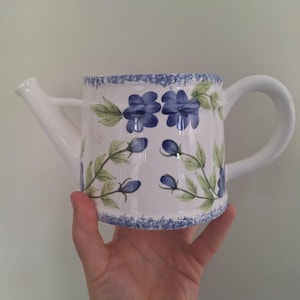 Op de afbeelding: Een witte keramische gieter met een handgeschilderd bloemmotief van blauwe bloemen, groene bladeren en blauwe knoppen. De rand en de basis zijn versierd met een blauwe gespikkelde rand, inclusief een gebogen handvat en een zijtuit.