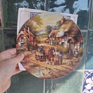 May include: A round decorative plate featuring a detailed pastoral scene of a horse-drawn cart on a dirt path between thatched-roof cottages. The illustration includes a person driving the cart, two people standing near the horse, a small dog, and several chickens in the foreground. The artwork is signed C.D. Vining.