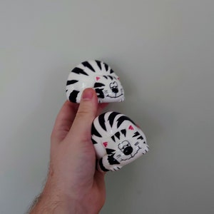 May include: Two ceramic cat figurines, painted in black and white stripes with pink accents. The cats are curled up in a sleeping position.